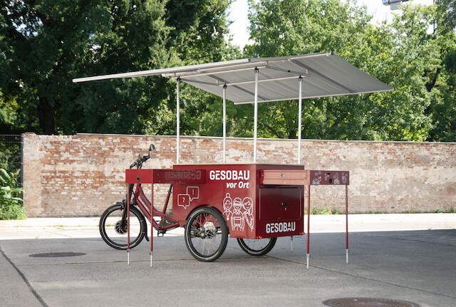 A bordeau red cargo bike from Radkutsche GmbH, called a Musketier, is showcased with a infostand, roof structure and retracktable drawers.
