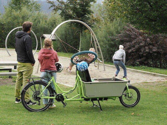 A father and mother stand besides there cargo bike while grandfather is playing with their childeren