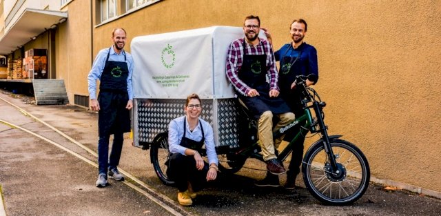 a team of 4 happily show of the 3 wheel cargo bike in front of a yellow orange wall