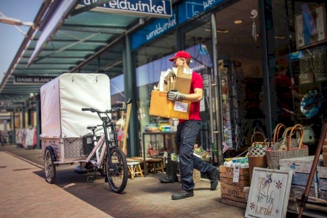 A garbage man comes out of a shop and walks to his three-wheeled bicycle, which he uses to collect rubbish.