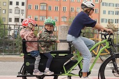 A mother rides her green cargo bike with two children on the back and laughs loudly.