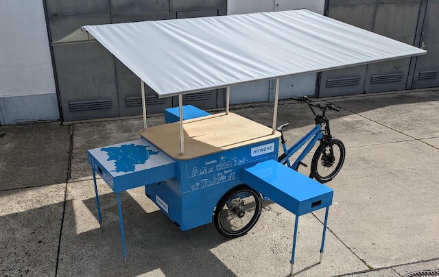 Ein blaues Lastenrad mit Informationsaufbau und grauer Dachkonstruktion wird in einer industriellen Betonumgebung gezeigt.