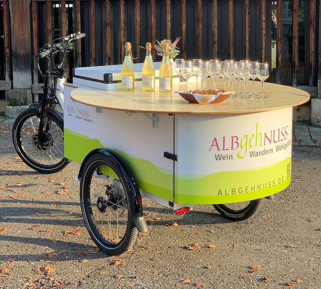 Ein Lastenrad mit drei Rädern und einem runden Tisch auf der Ladefläche wird verwendet, um Kunden in einer natürlichen Umgebung mit Wein oder Sekt zu versorgen.